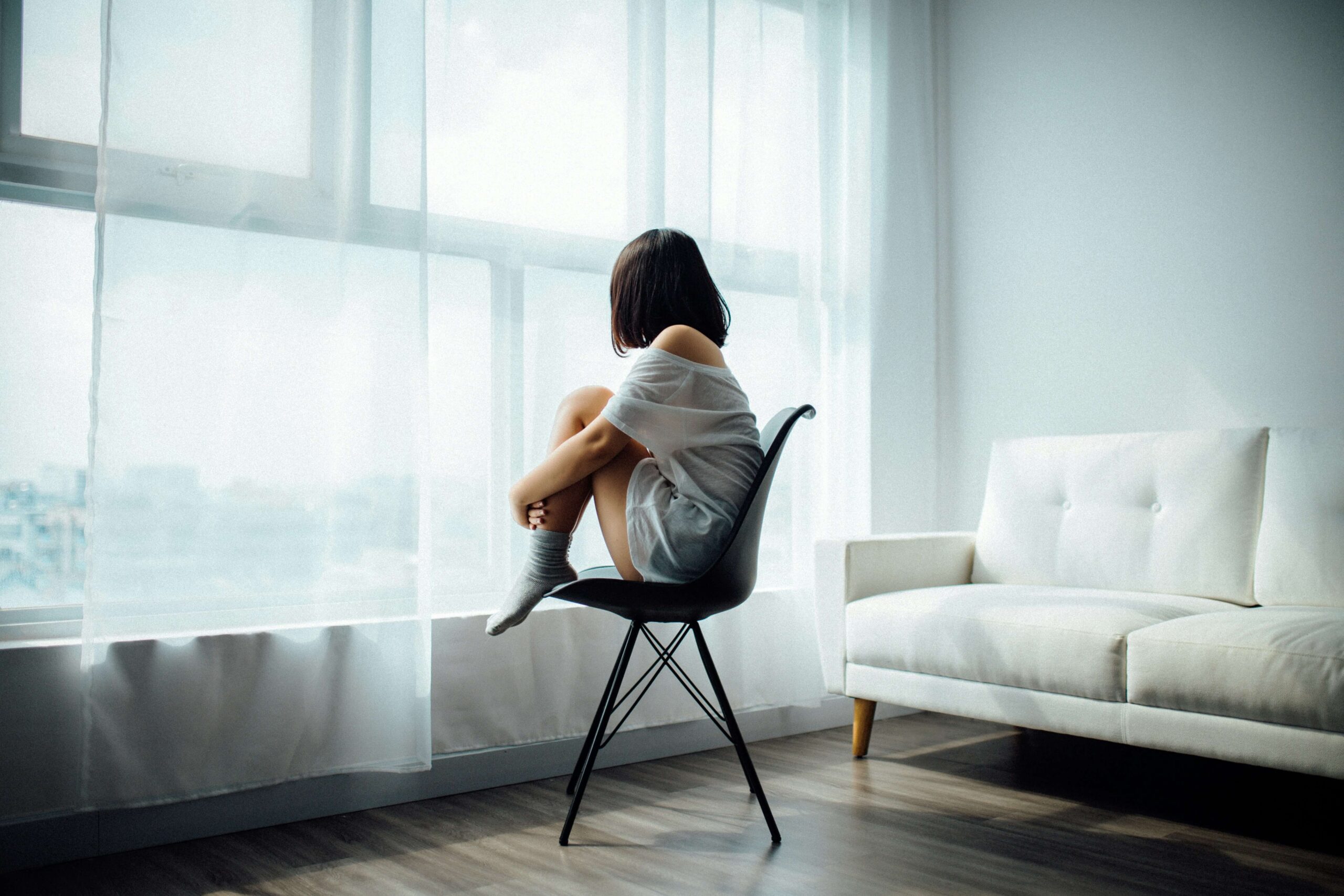 child sat on chair with feet up hugging knees and looking out of the window